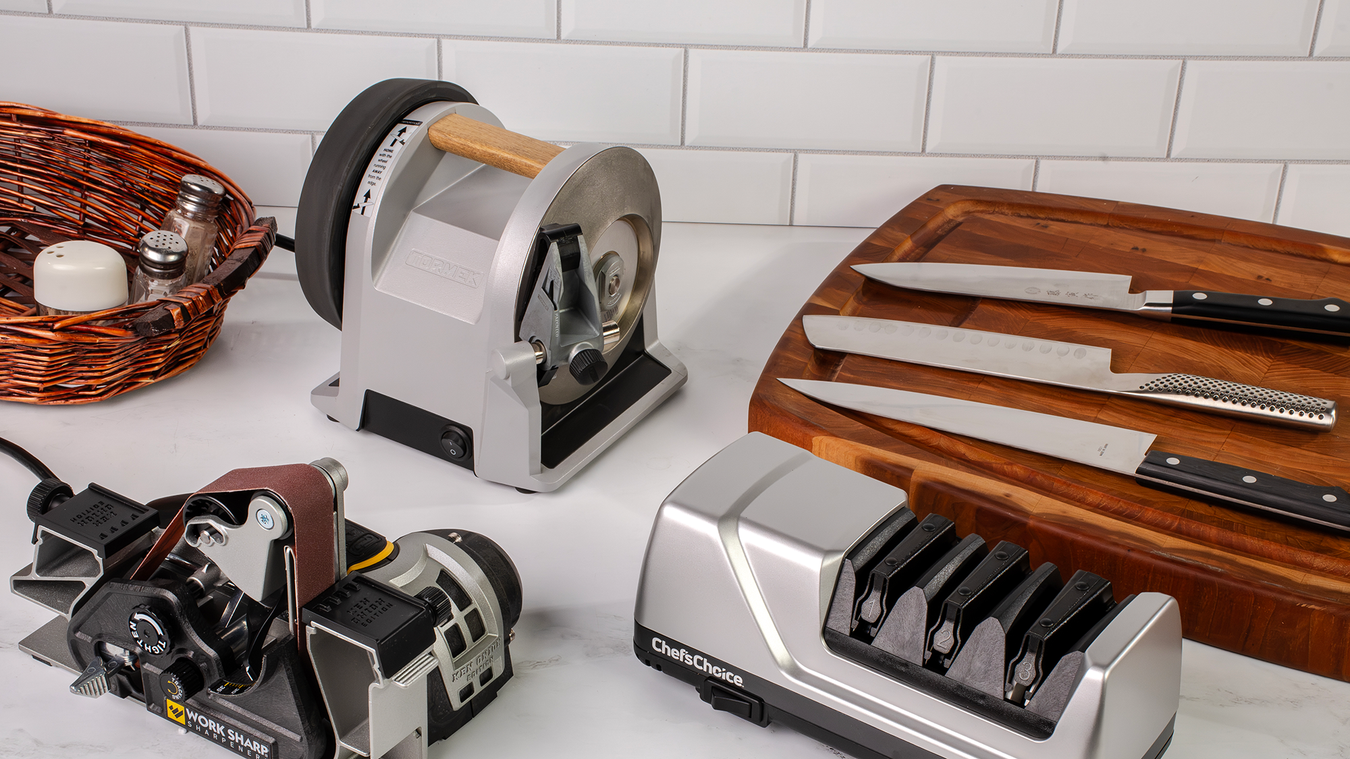 A set of kitchen knife sharpening tools arranged on a white countertop against a white subway tile backsplash, alongside knives on a wooden cutting board.